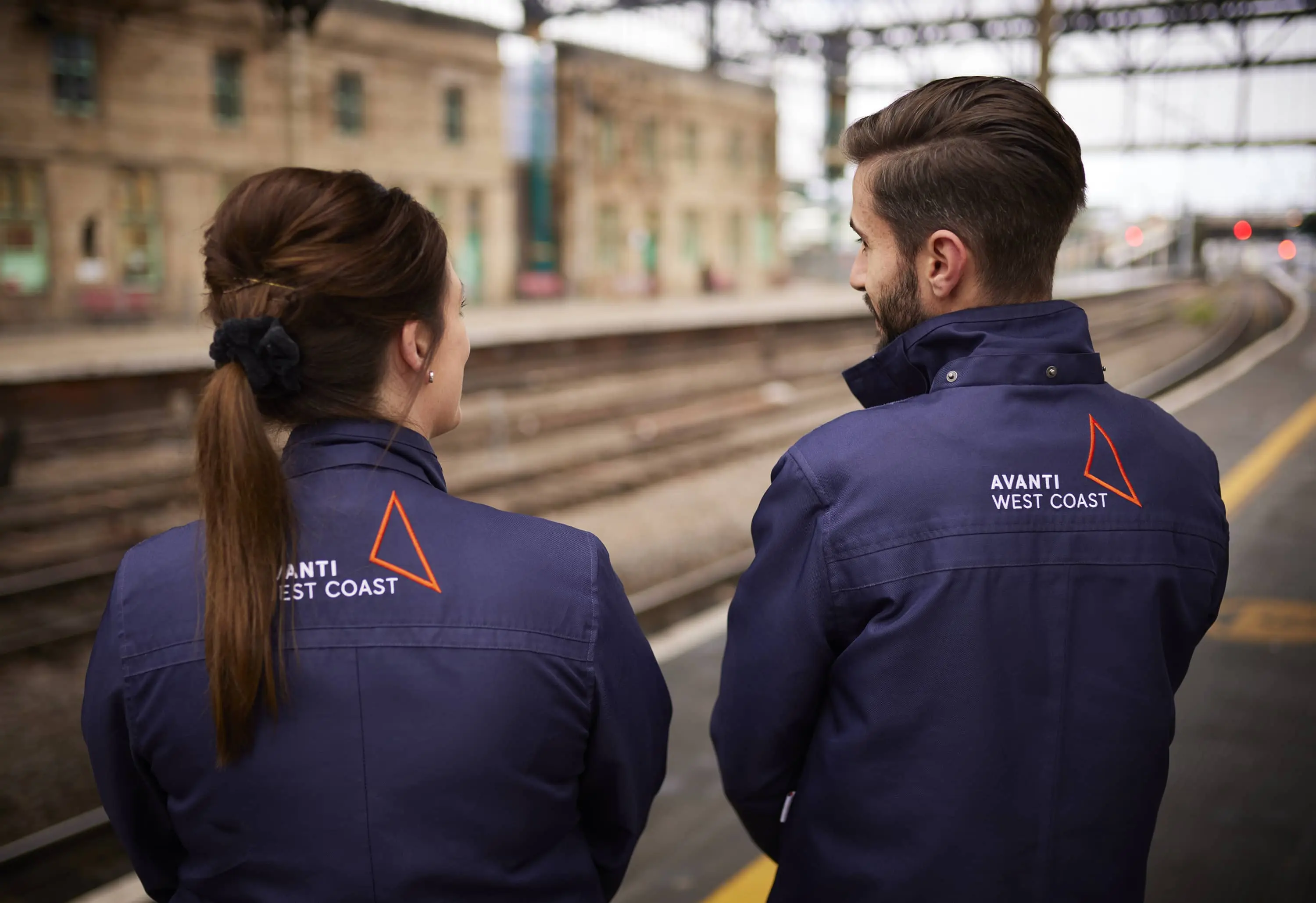 Two staff members in navy Avanti jackets stand on a station platform, looking out towards the railway tracks.